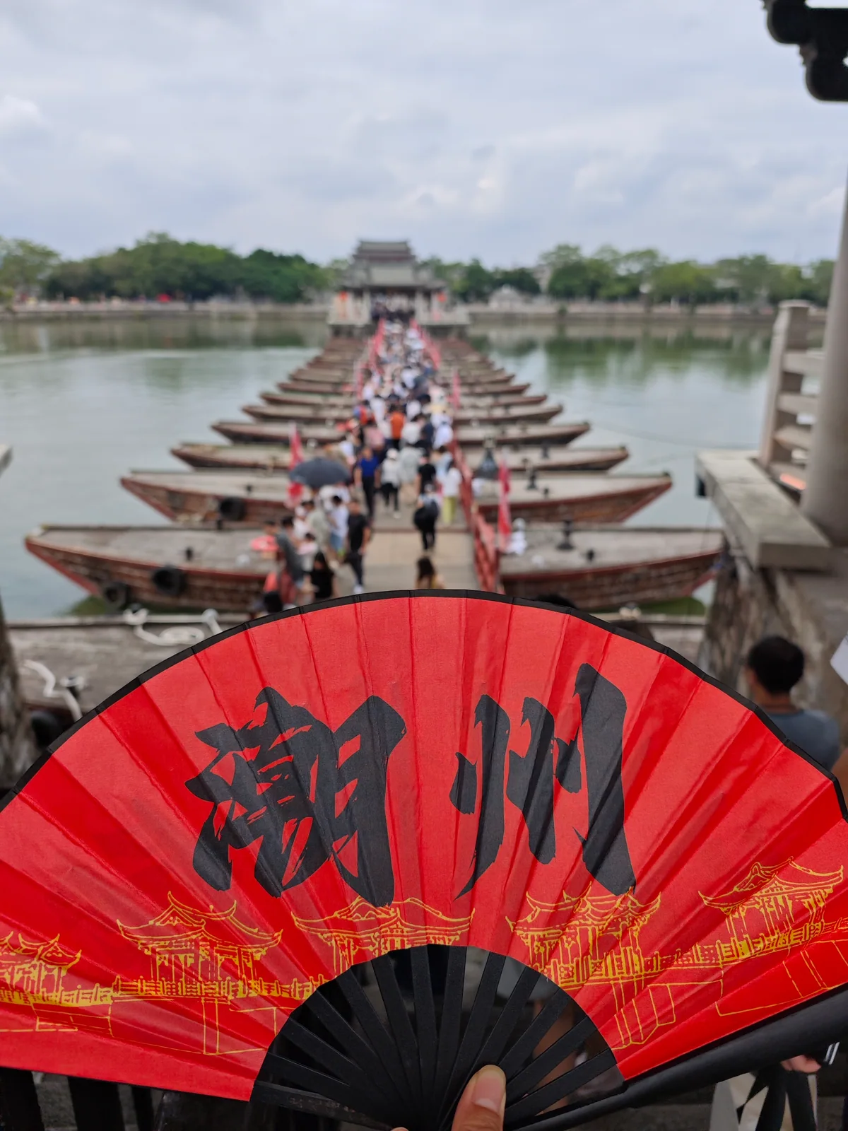 A red folding fan marked Chaozhou in front of the wooden boats and crowded walkway of Guangji Bridge / 写着“潮州”的红色折扇前景，背景是广济桥的梭船浮桥与过桥人群