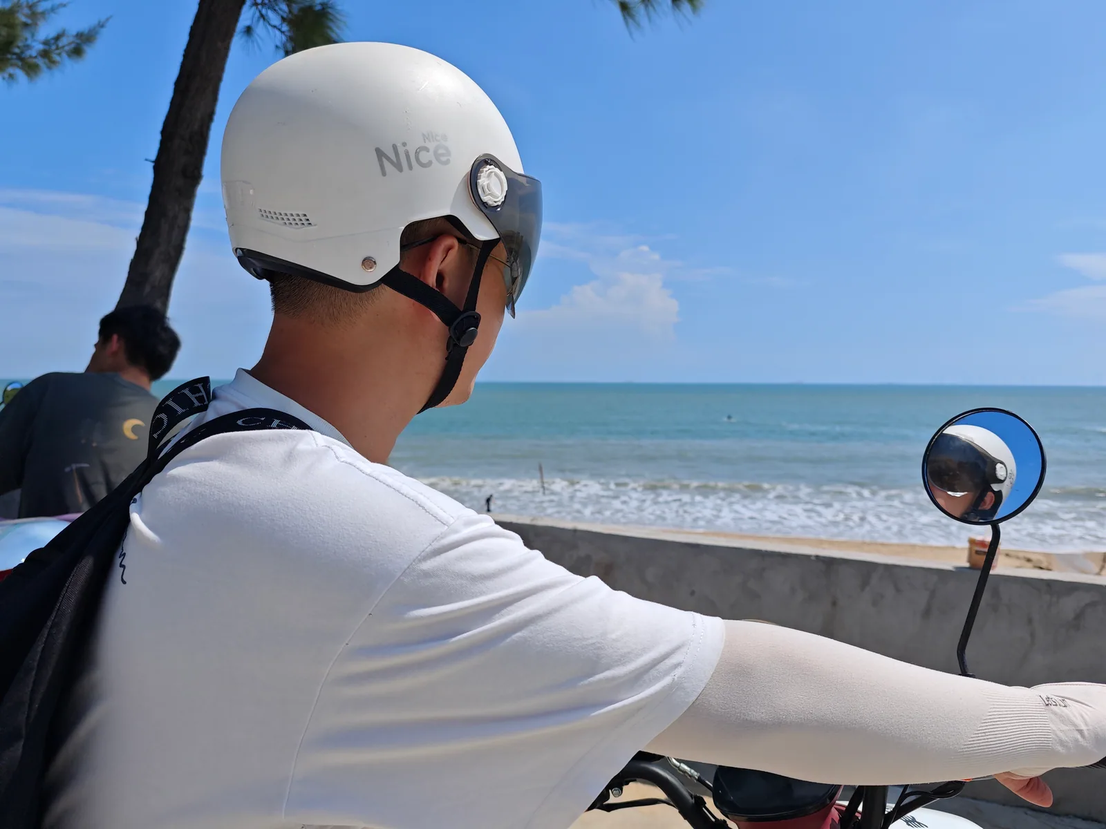 A rider wearing a white helmet beside the sea, with the helmet reflected in the rear-view mirror / 戴白色头盔的骑行者身旁是海面，后视镜中映着头盔