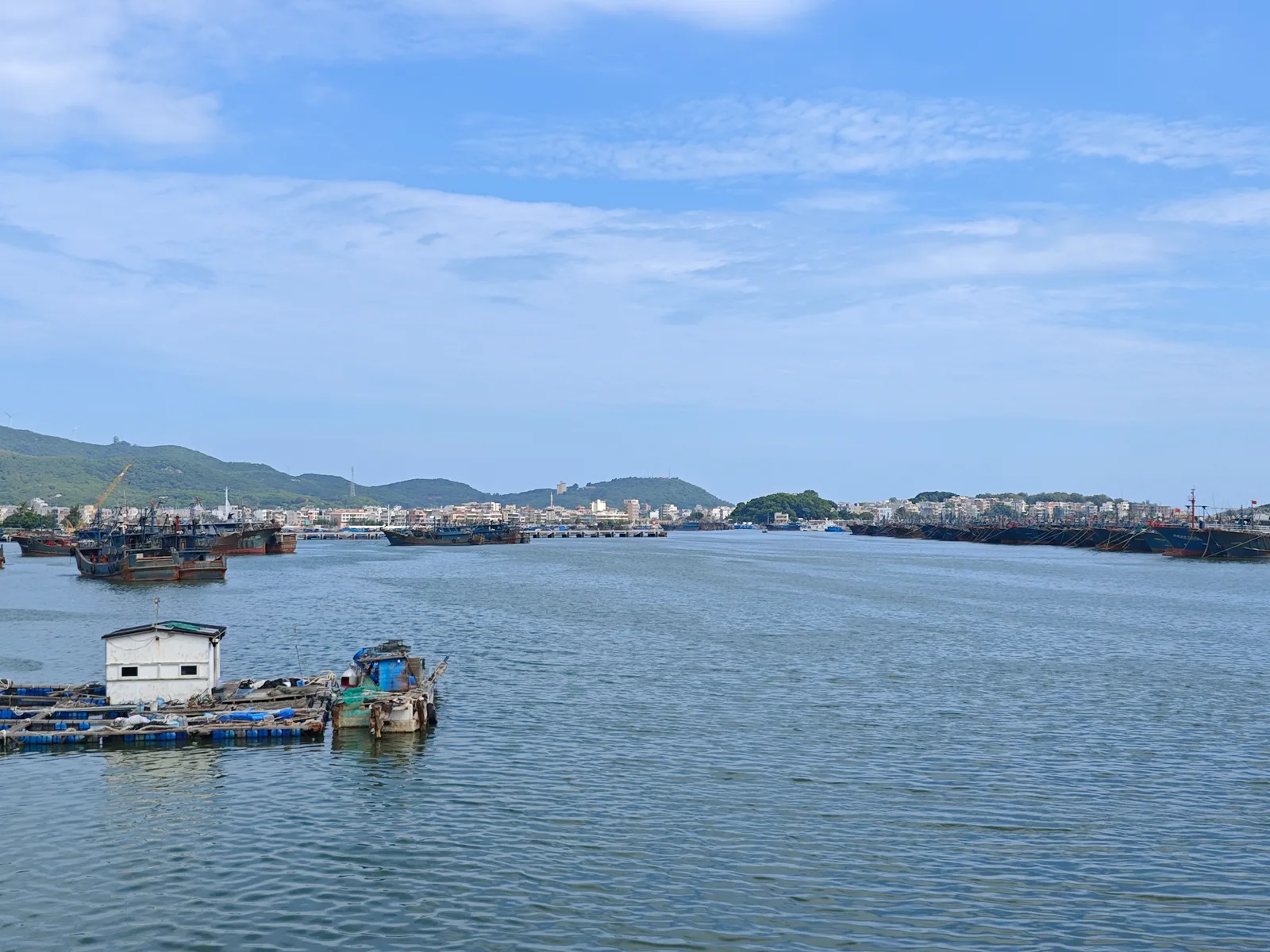 Fishing boats moored across a blue harbour, with floating platforms in front and a town below green hills / 蓝色渔港内成排停泊的渔船，近处有浮排，远处是山下的城镇
