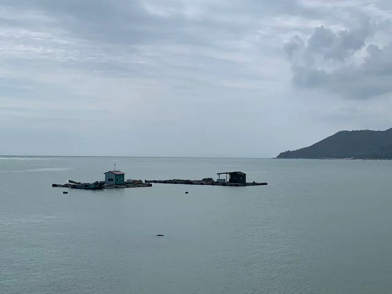 Two floating platforms with small huts on a calm grey-blue sea, with a headland to the right / 灰蓝色平静海面上的两处浮排与小屋，右侧是一段山岬