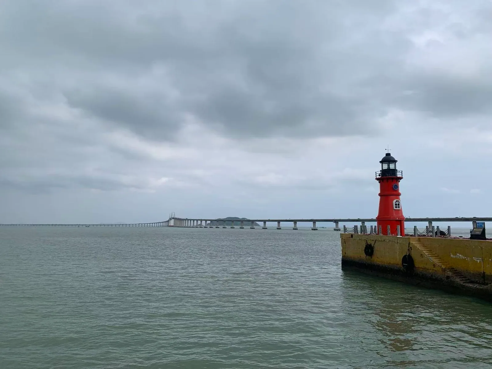 A red lighthouse on a pier, with a long sea bridge beneath a cloudy sky / 阴云下的红色灯塔立在码头边，远处跨海大桥横过海面