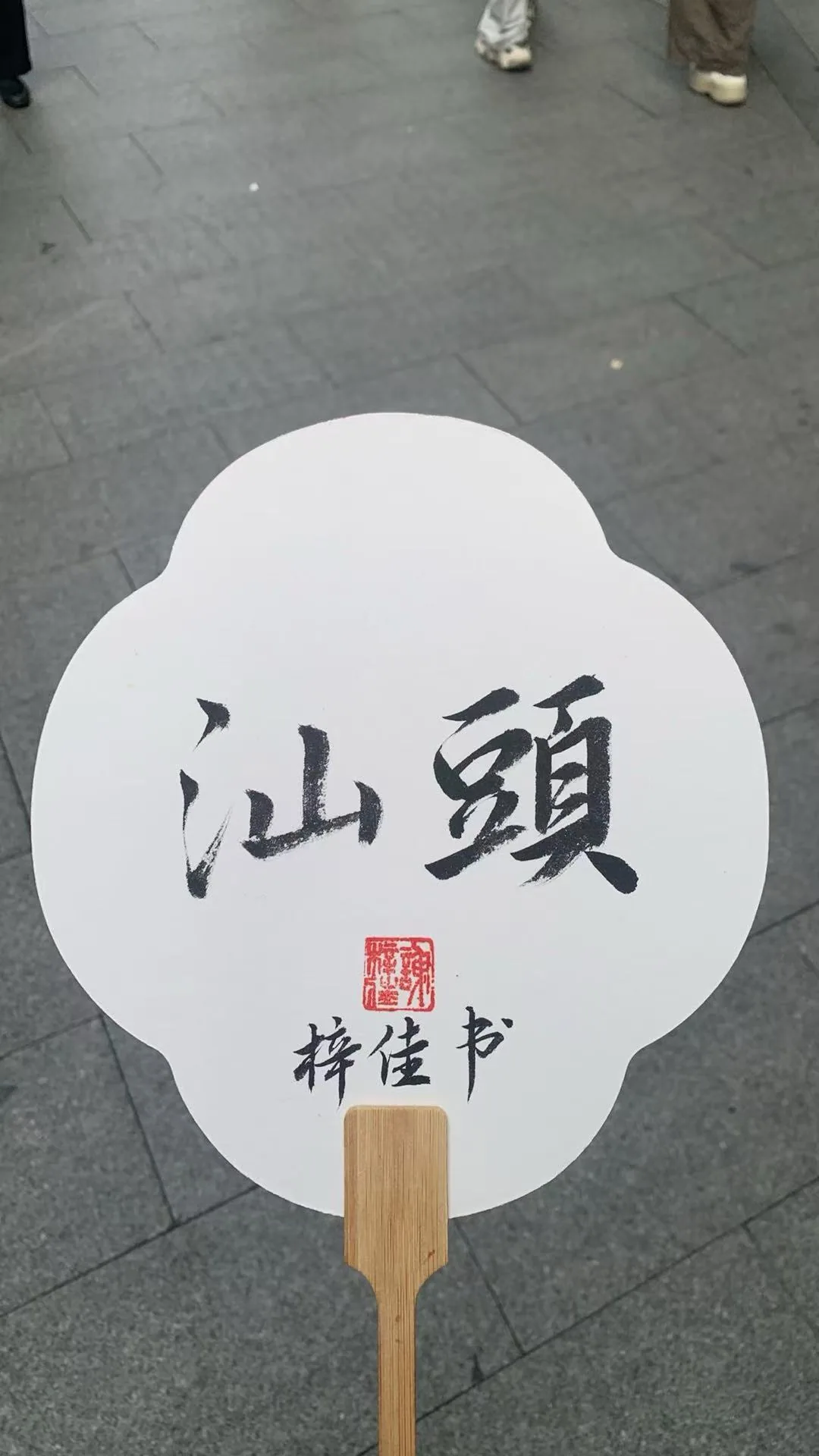 A white handheld fan bearing the brush-written characters for Shantou, against grey paving / 灰色石板路前，一把写着“汕頭”的白色手持团扇